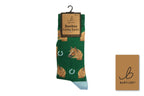 A pair of green Bartleby Bamboo Funky Socks hanging on a black hanger, featuring a pattern of brown coiled shapes, white horseshoes, and a horse head. A small brown brand card is shown to the right on a white background.