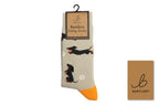 A pair of Bartleby Bamboo Funky Socks featuring a beige base with black and brown dachshund illustrations, white paw prints, and bright orange toes, shown on a hanger with a brand card.