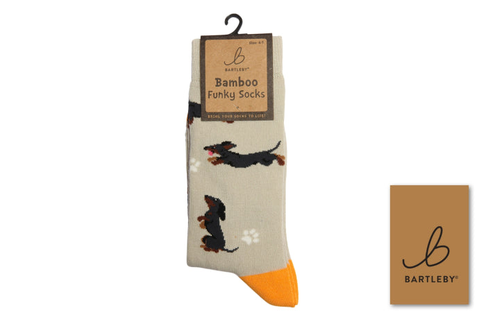 A pair of Bartleby Bamboo Funky Socks featuring a beige base with black and brown dachshund illustrations, white paw prints, and bright orange toes, shown on a hanger with a brand card.